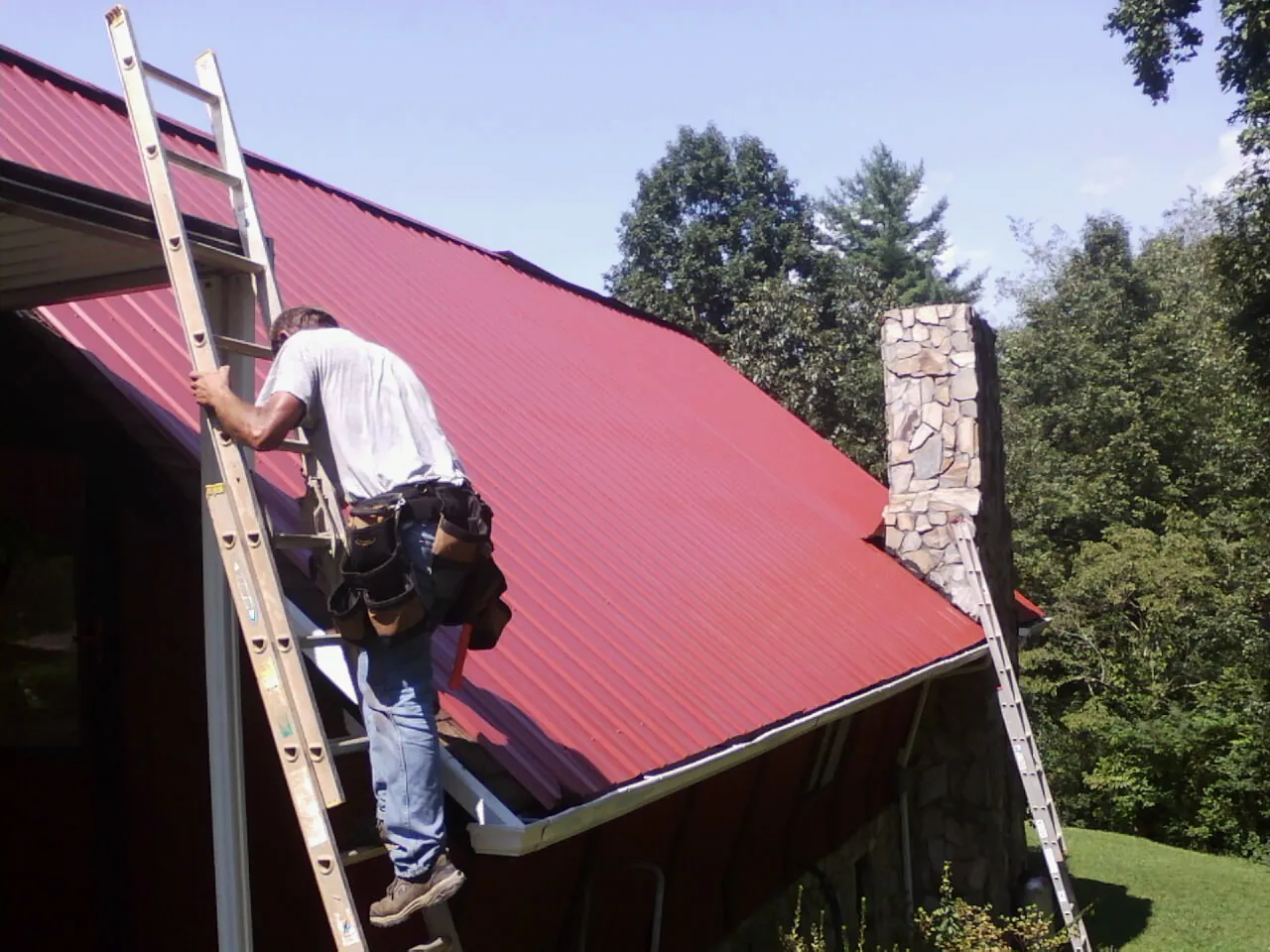 Roofer on a ladder beside a red metal roof and stone chimney — Soffit & Fascia Repair by Roofing Brothers in Rhododendron